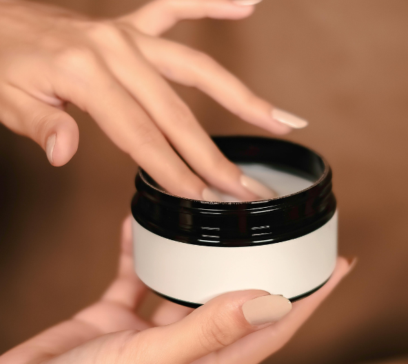 Mains touchant une crème dans un pot. Peau claire, ongles nude, produit dans un contenant blanc aux touches noires.