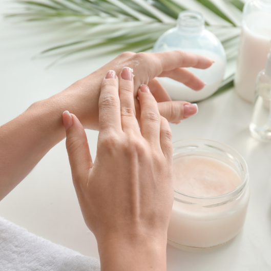 Des mains appliquent de la crème à partir d'un pot. D'autres produits de soin sont disposés sur une surface blanche.