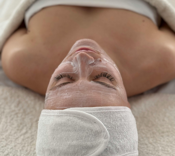 Femme avec de la crème pour le visage, les yeux fermés, portant un bandeau blanc et une serviette, au spa.