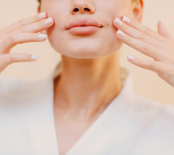 Femme touchant ses joues, gros plan, dans une robe blanche, avec une peau pâle et un fond de couleur claire.