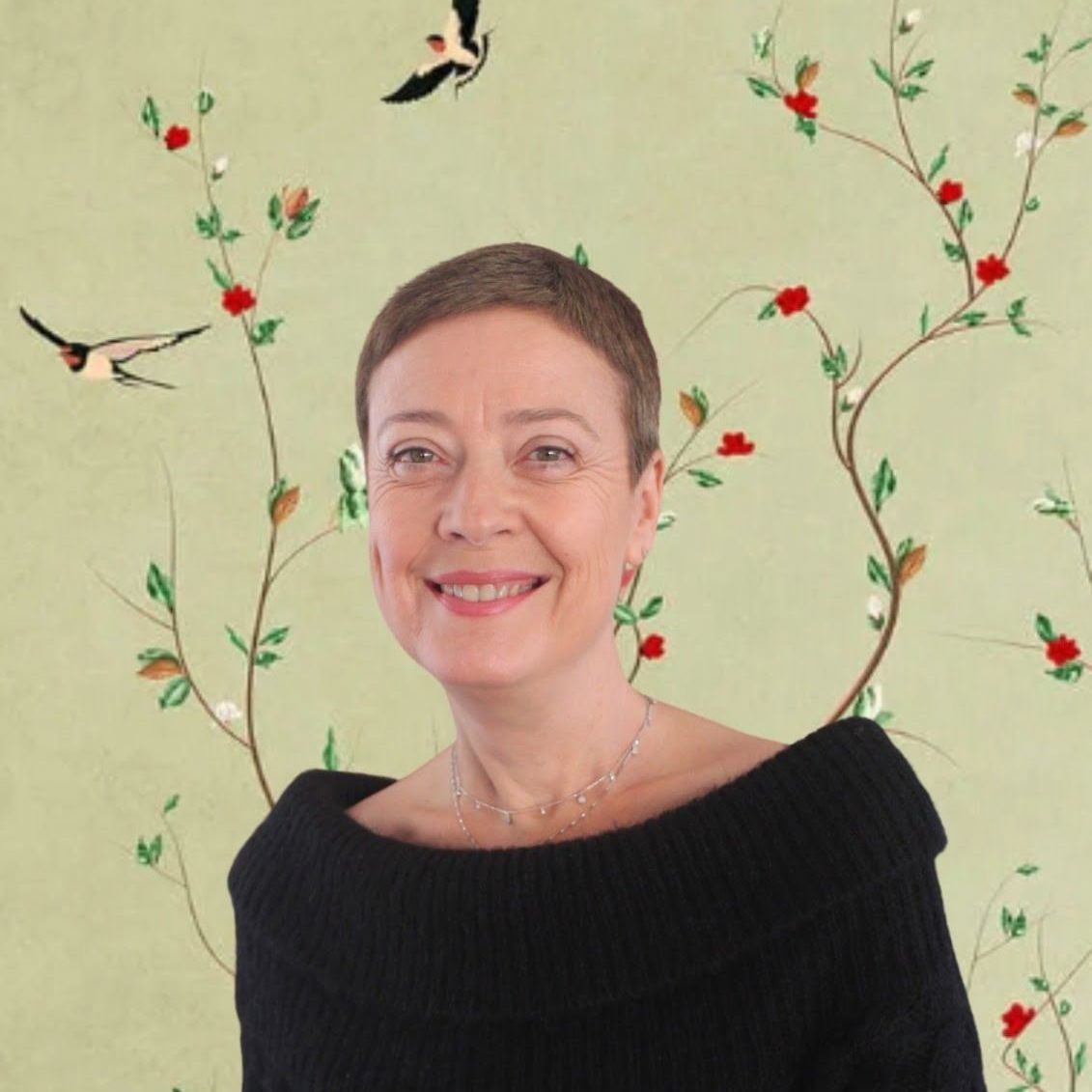 Une femme aux cheveux courts sourit, portant un pull noir, devant un papier peint floral vert.