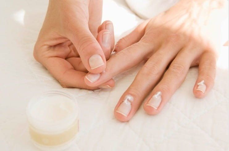 Mains appliquant de la crème sur les ongles à côté d'un pot de crème sur une surface blanche.