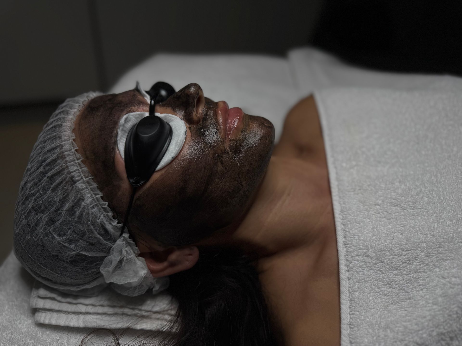 Eine Person mit Gesichtsmaske und Schutzbrille liegt auf einer Spa-Liege, um den Kopf ist ein Handtuch gewickelt.