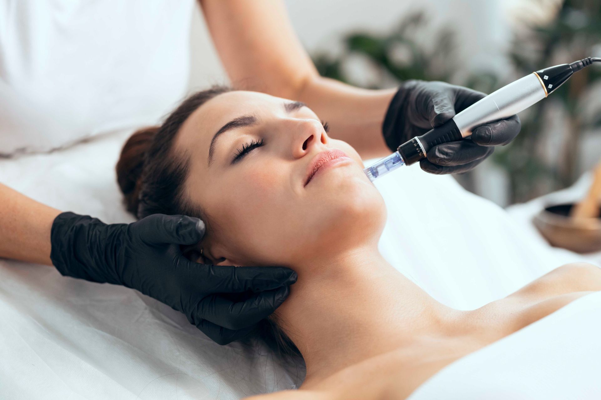 Eine Kosmetikerin führt eine Gesichtsbehandlung mit einem Handgerät an einer liegenden Kundin auf einer Spa-Liege durch.