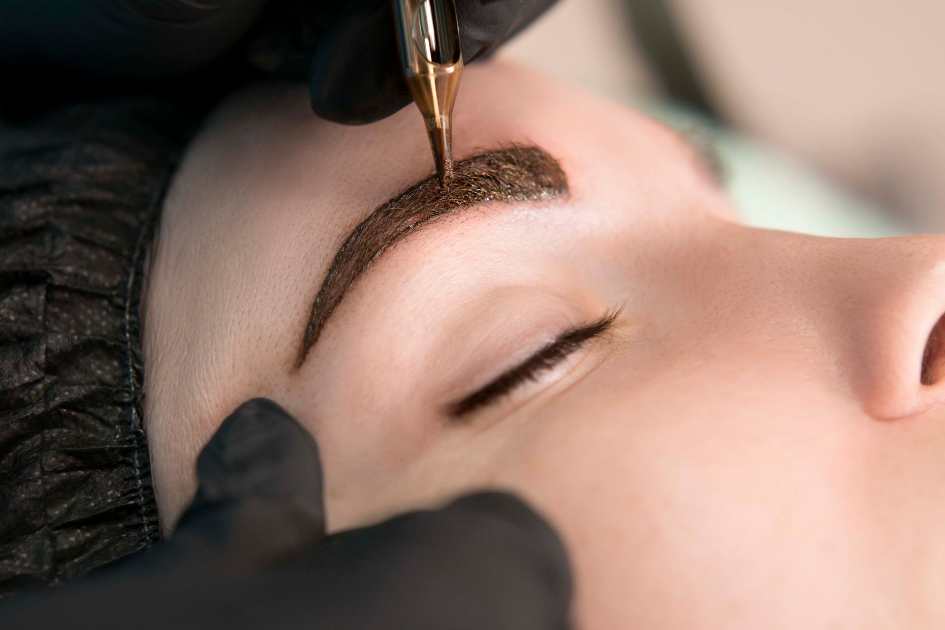 Kosmetikerin trägt Augenbrauen-Make-up mit einem Pinsel auf das geschlossene Auge einer Kundin auf