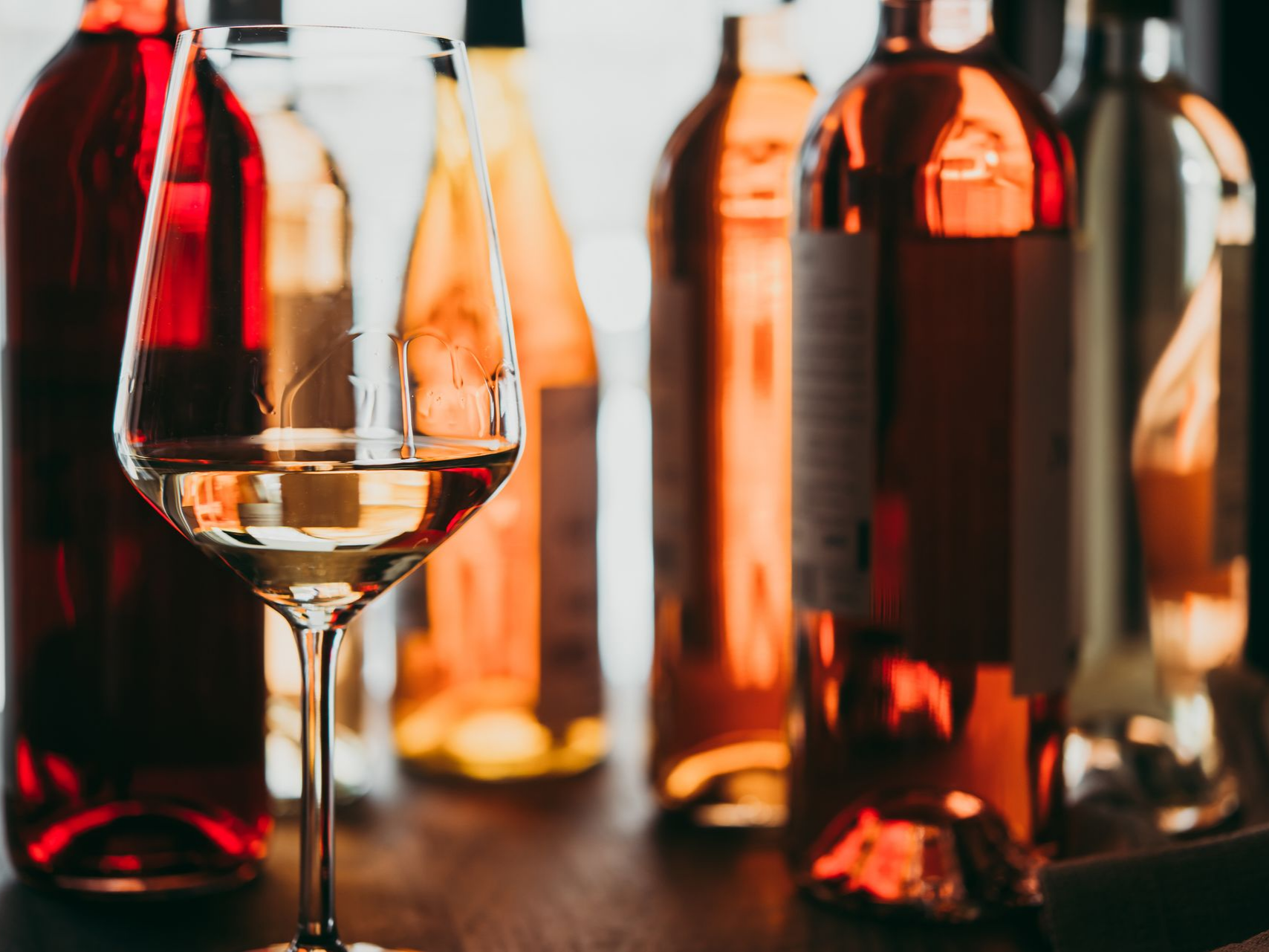 Verre de vin clair au premier plan, bouteilles de vin de couleurs variées en arrière-plan.