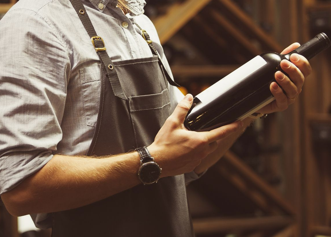 Un homme tenant une bouteille de vin, portant un tablier, dans une cave à vin.