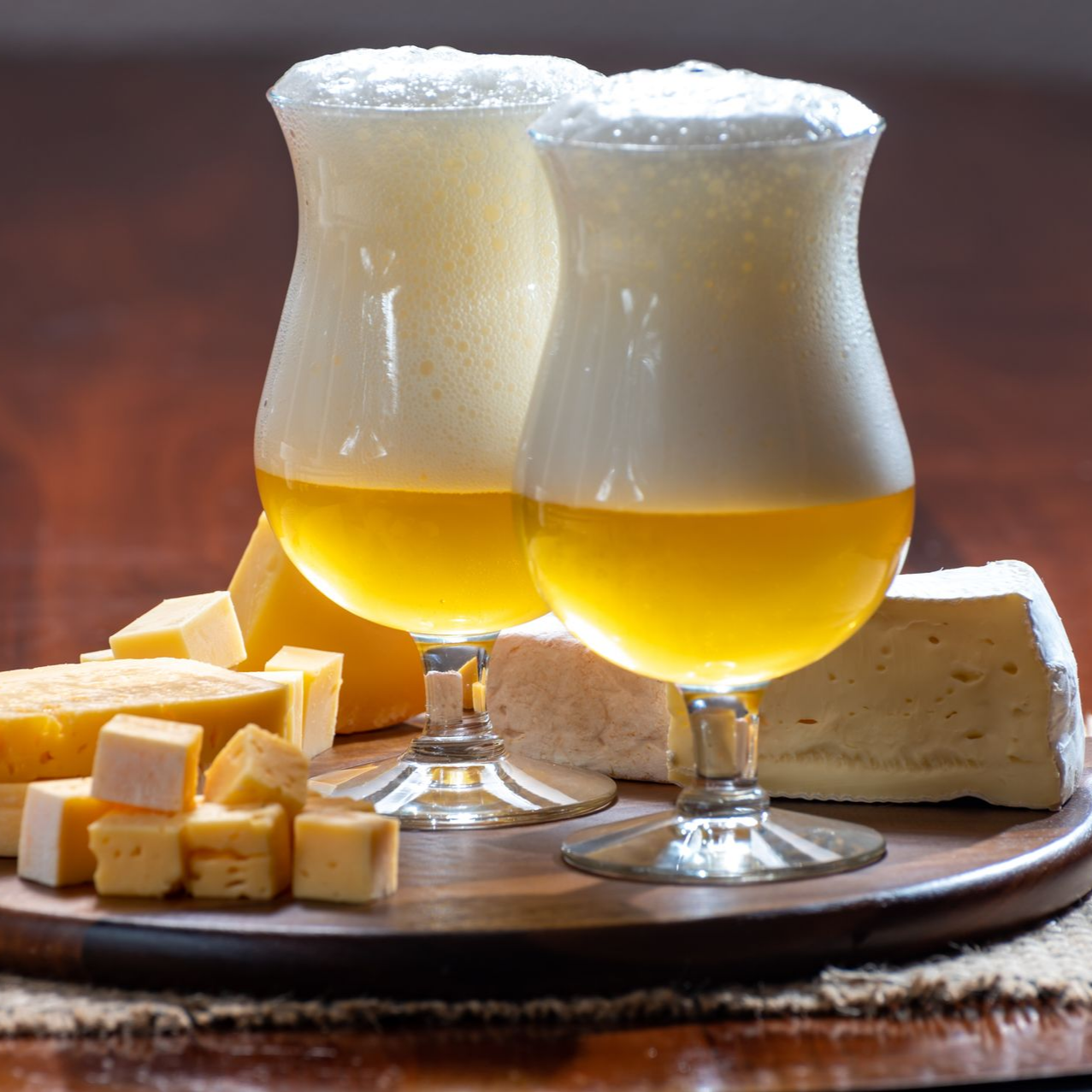 Deux verres de bière à la mousse onctueuse et du fromage sur un plateau en bois.
