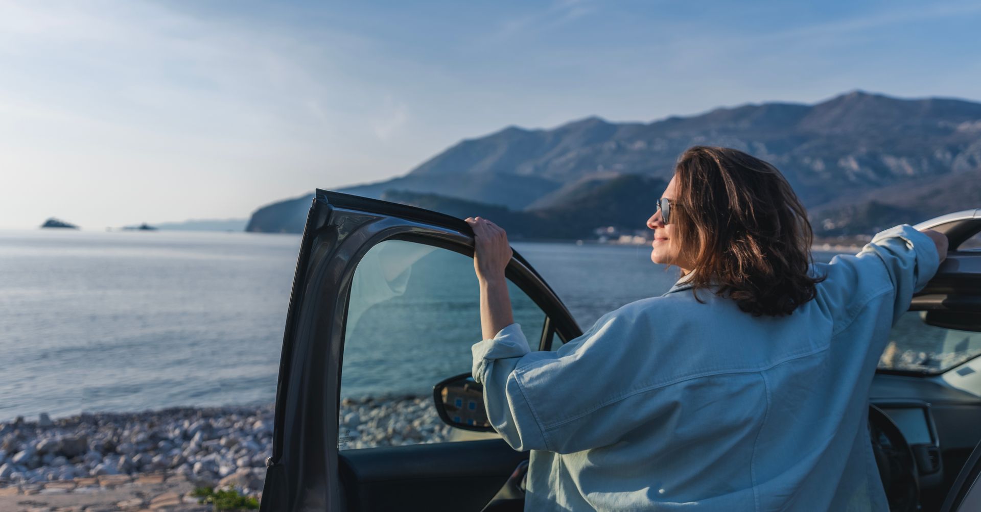 Une femme portant des lunettes de soleil est appuyée contre la portière d'une voiture, profitant de la vue sur l'océan et les montagnes.