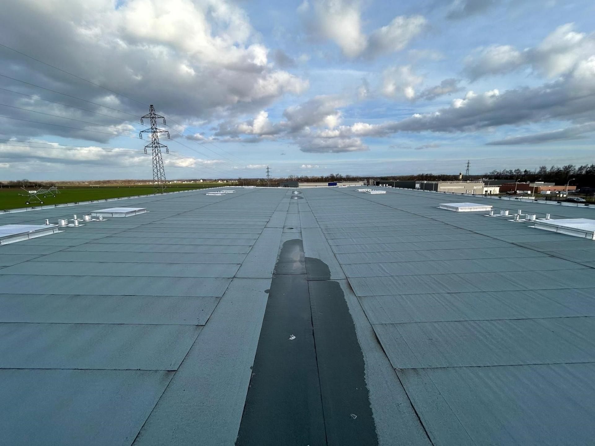 Toit plat et gris, d'origine commerciale, sous un ciel légèrement nuageux, avec des lignes électriques en arrière-plan.