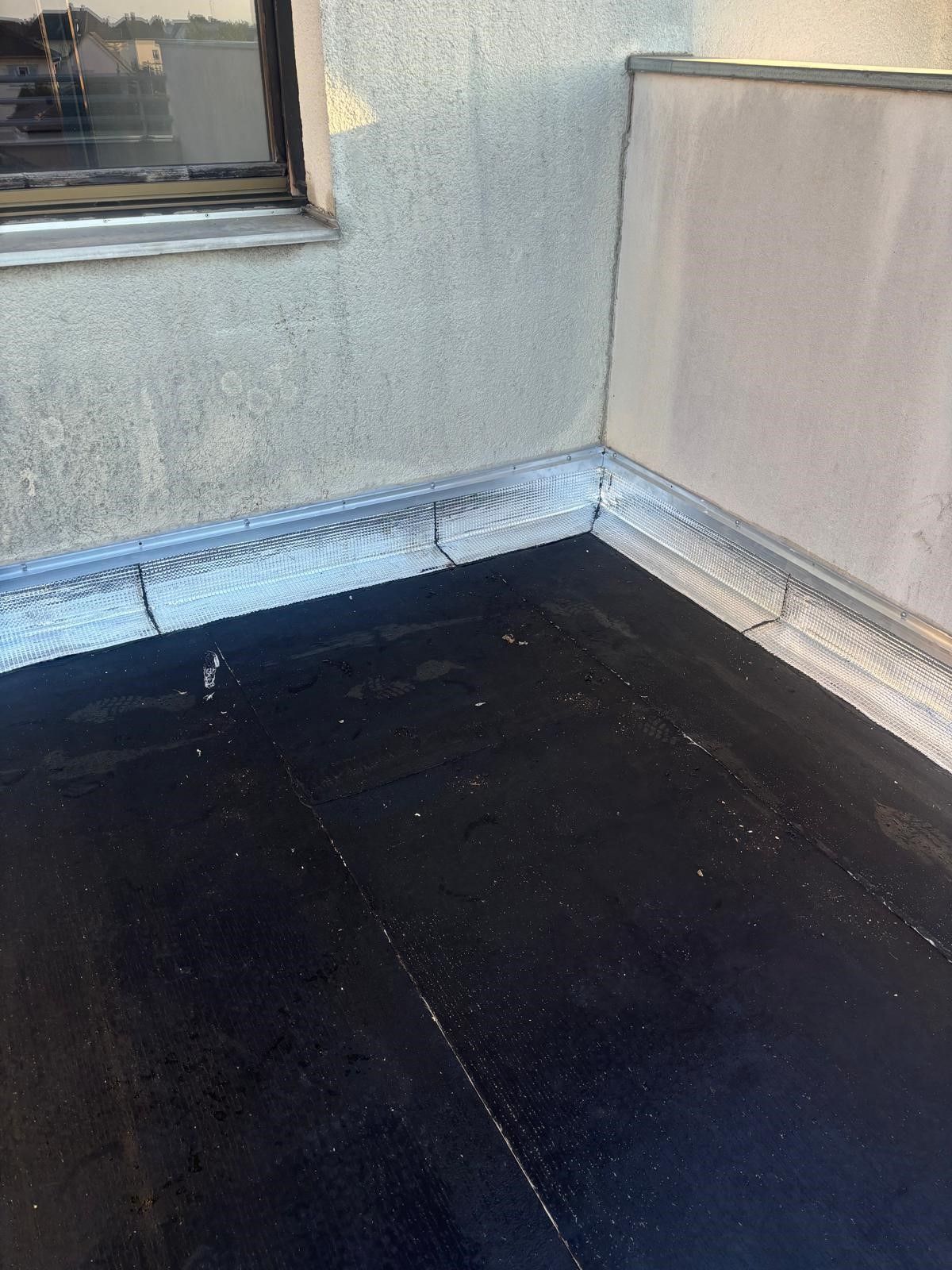 Toit plat noir avec bordure à l'angle d'un bâtiment, à côté d'une fenêtre et d'un mur.