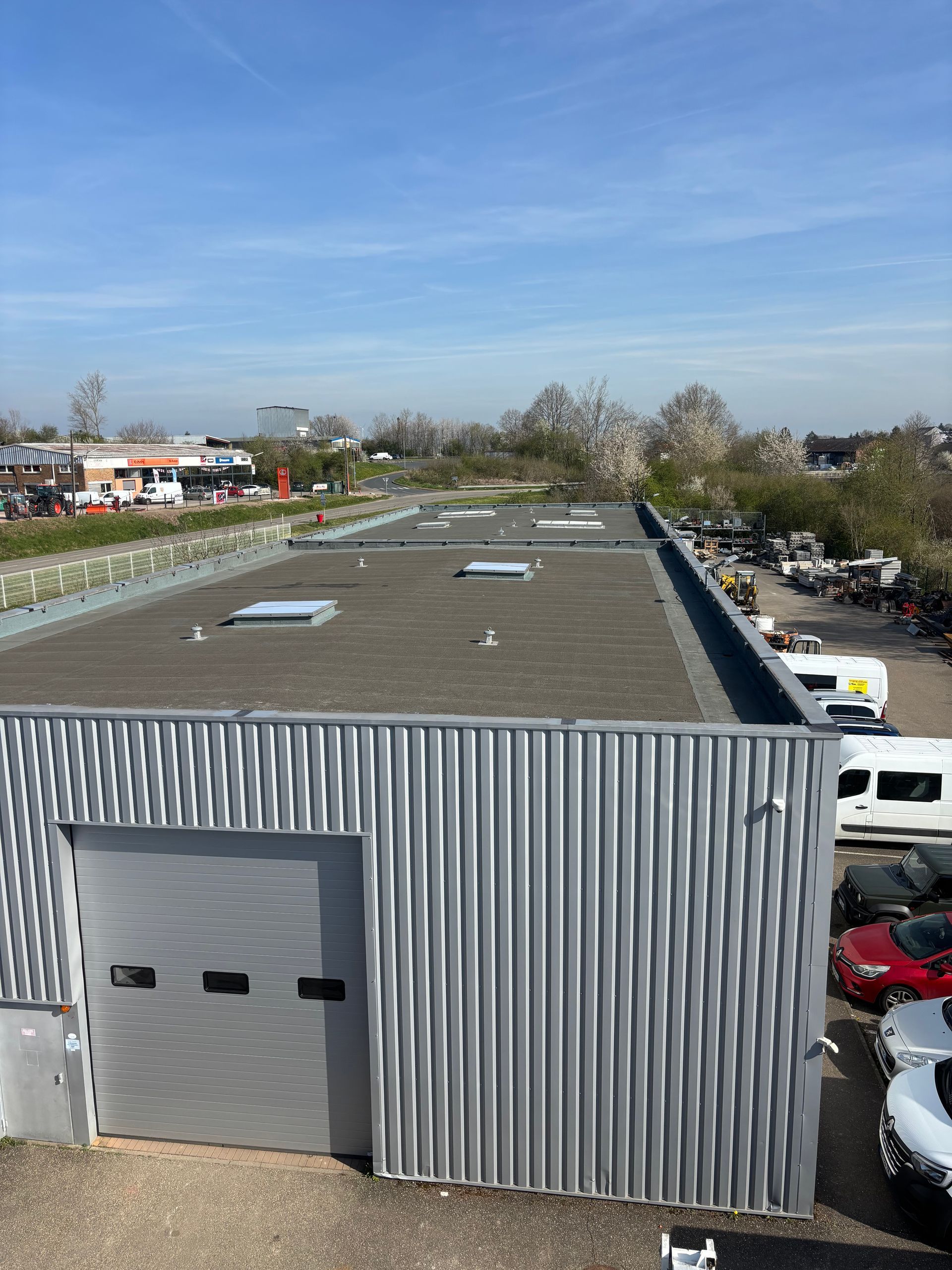 Un bâtiment en tôle ondulée à toit plat se dresse sous un ciel bleu. Des véhicules et des arbres apparaissent en arrière-plan.