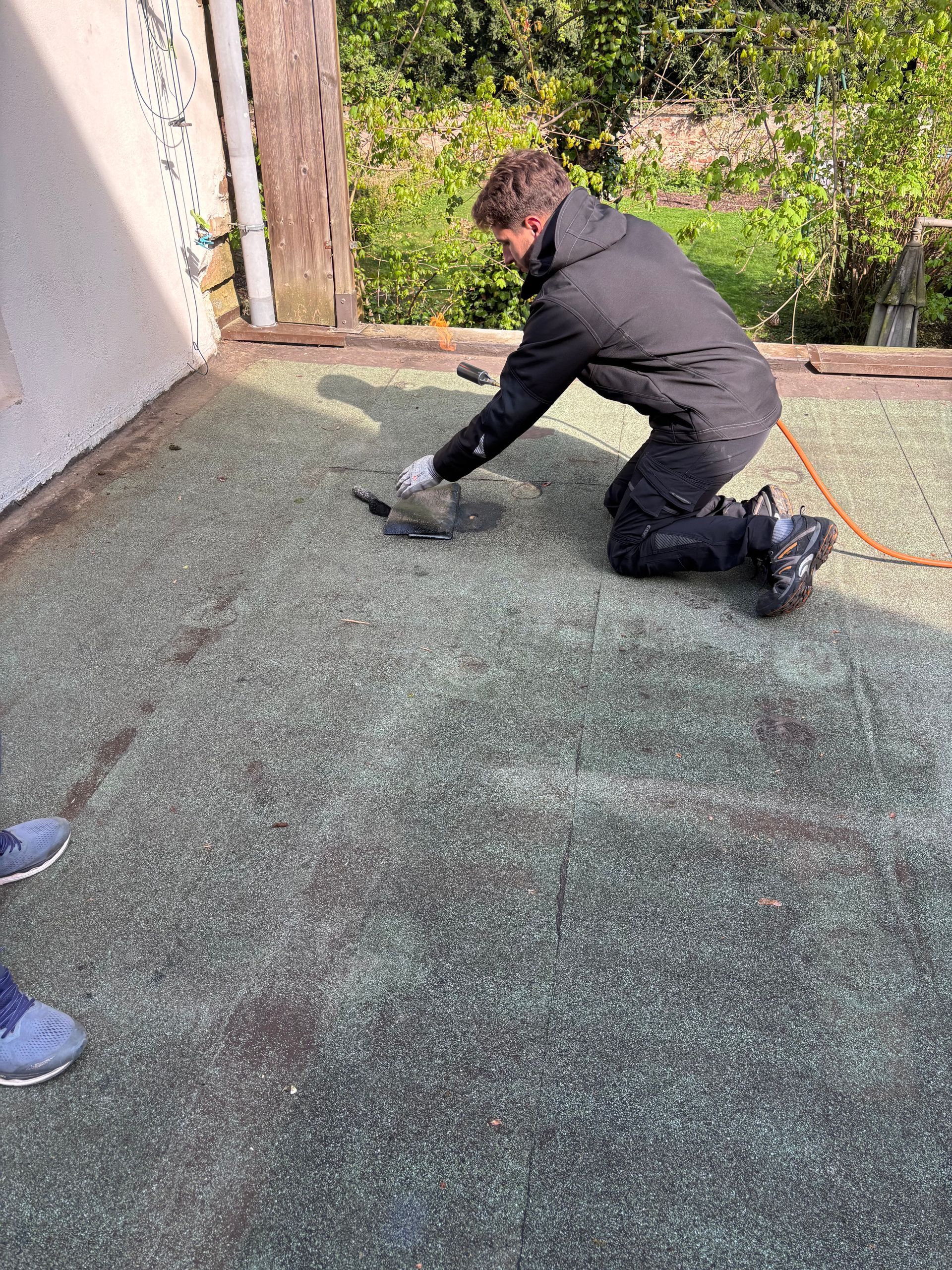 Personne agenouillée sur un toit plat, travaillant avec un outil. Matériau de toiture vert et noir. Extérieur.