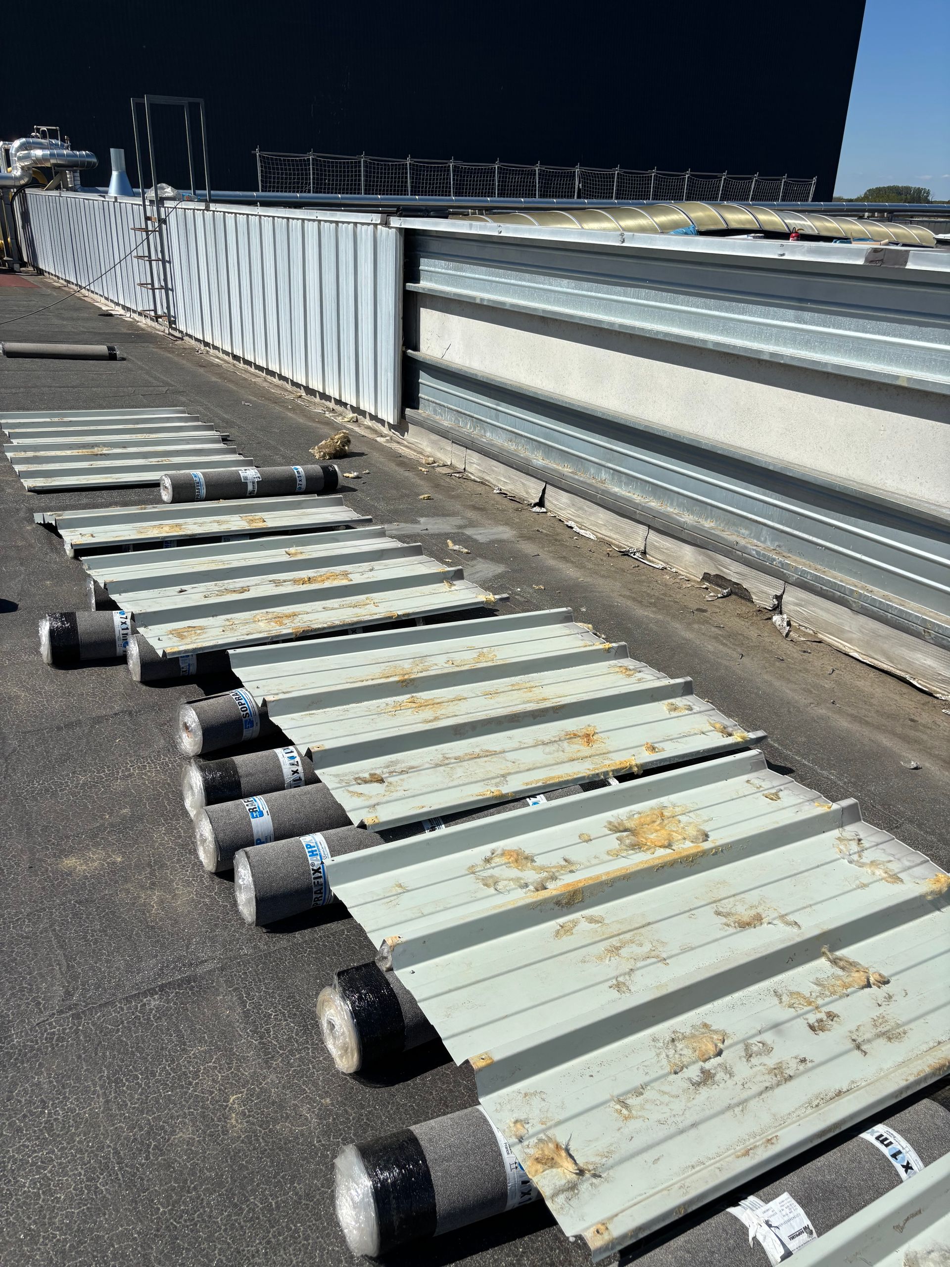 Des panneaux de toiture métalliques et des rouleaux de matériau sont alignés sur l'asphalte à côté d'un bâtiment.