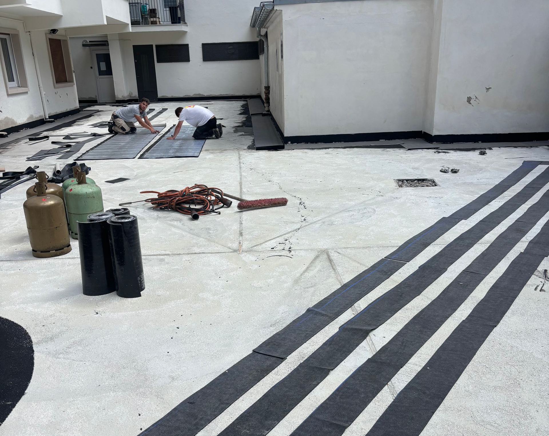 Des ouvriers posent une toiture sur un bâtiment ; rouleaux d’asphalte, chalumeaux et bandes finies sur une surface en béton.