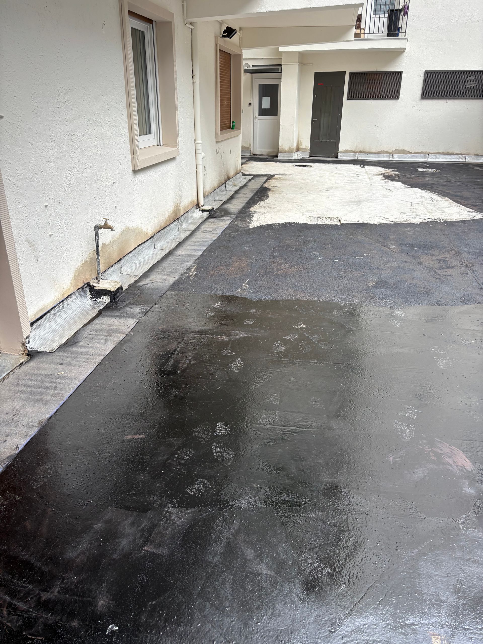 Allée en asphalte sombre et mouillée, avec des flaques d'eau, près d'un bâtiment. Murs blancs, fenêtre et porte visibles.