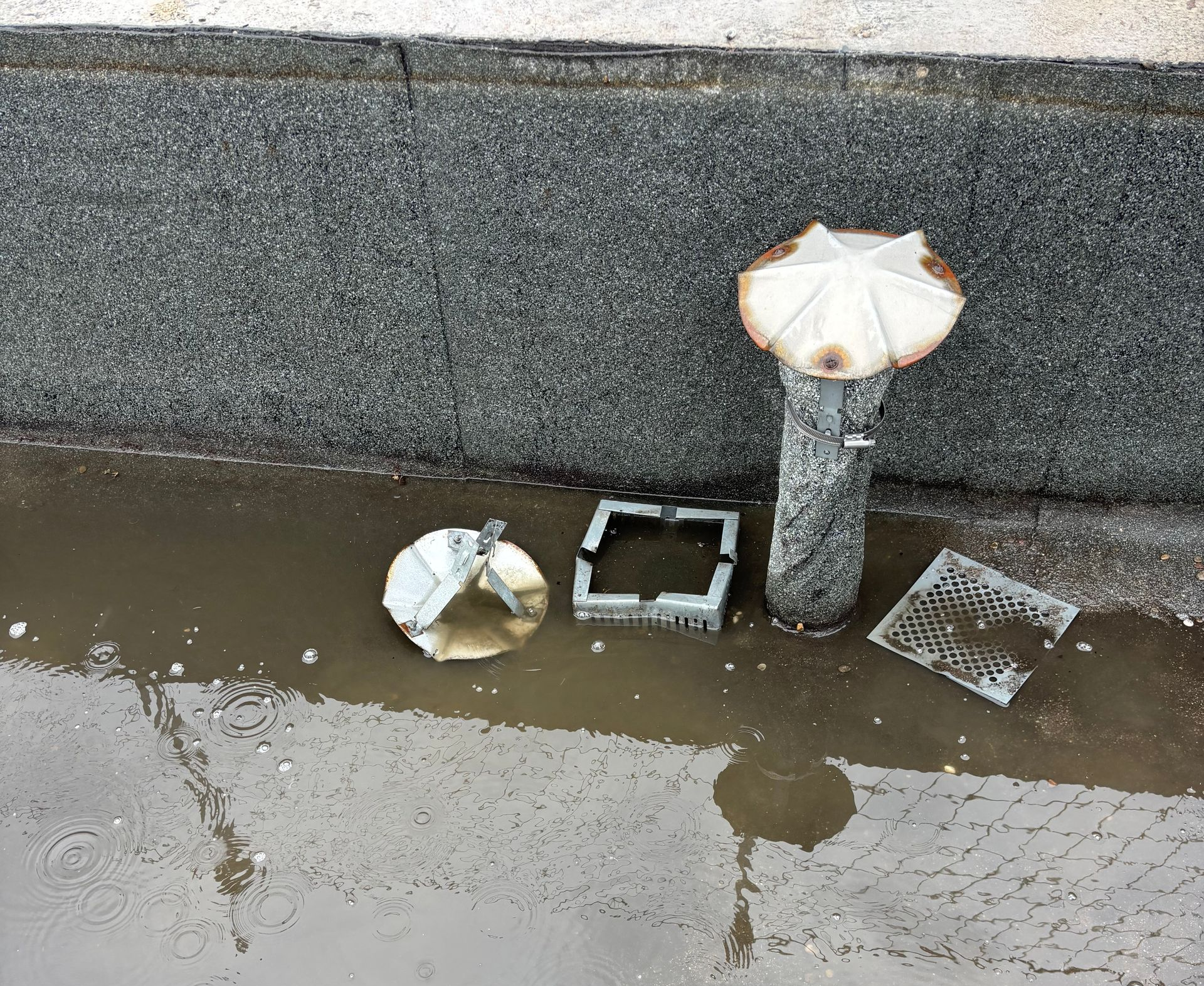 Une flaque d'eau se trouve à côté d'un mur texturé gris foncé, avec des éléments de canalisation d'eaux pluviales éparpillés dans l'eau.