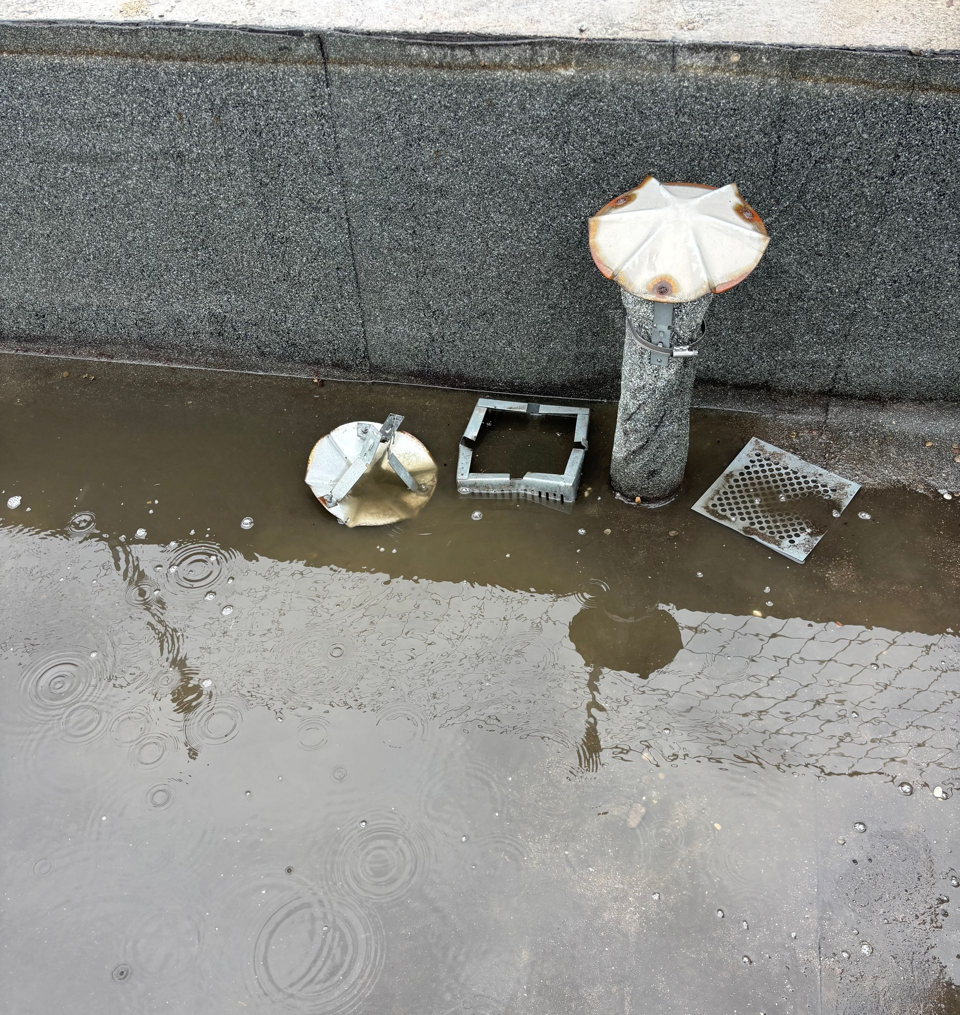 Un drain d'eau pluviale dont les grilles ont été retirées et une flaque d'eau stagnante sur un toit.