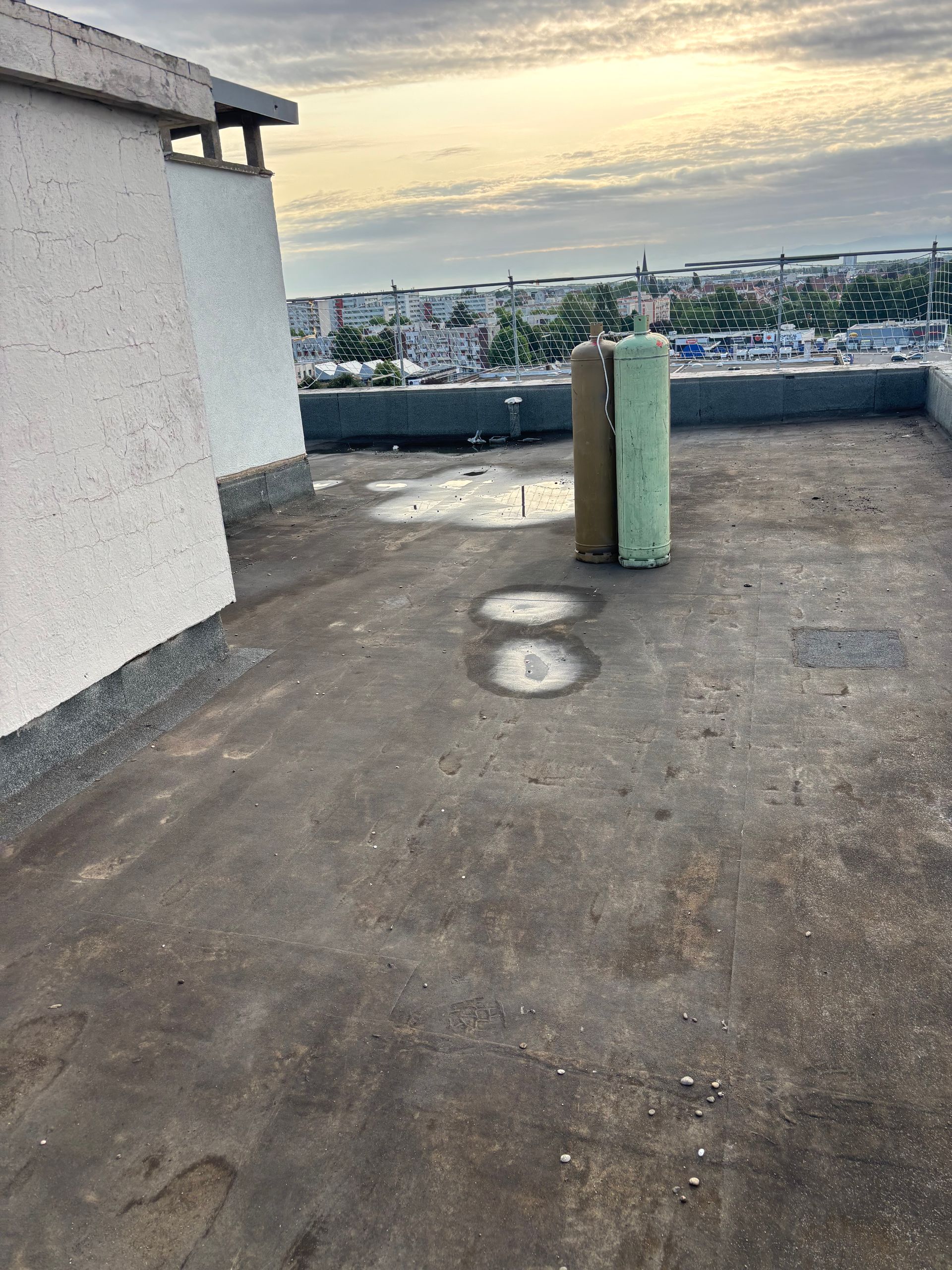 Toit avec deux objets cylindriques, des traces d'eau et une ville en arrière-plan sous un ciel nuageux.