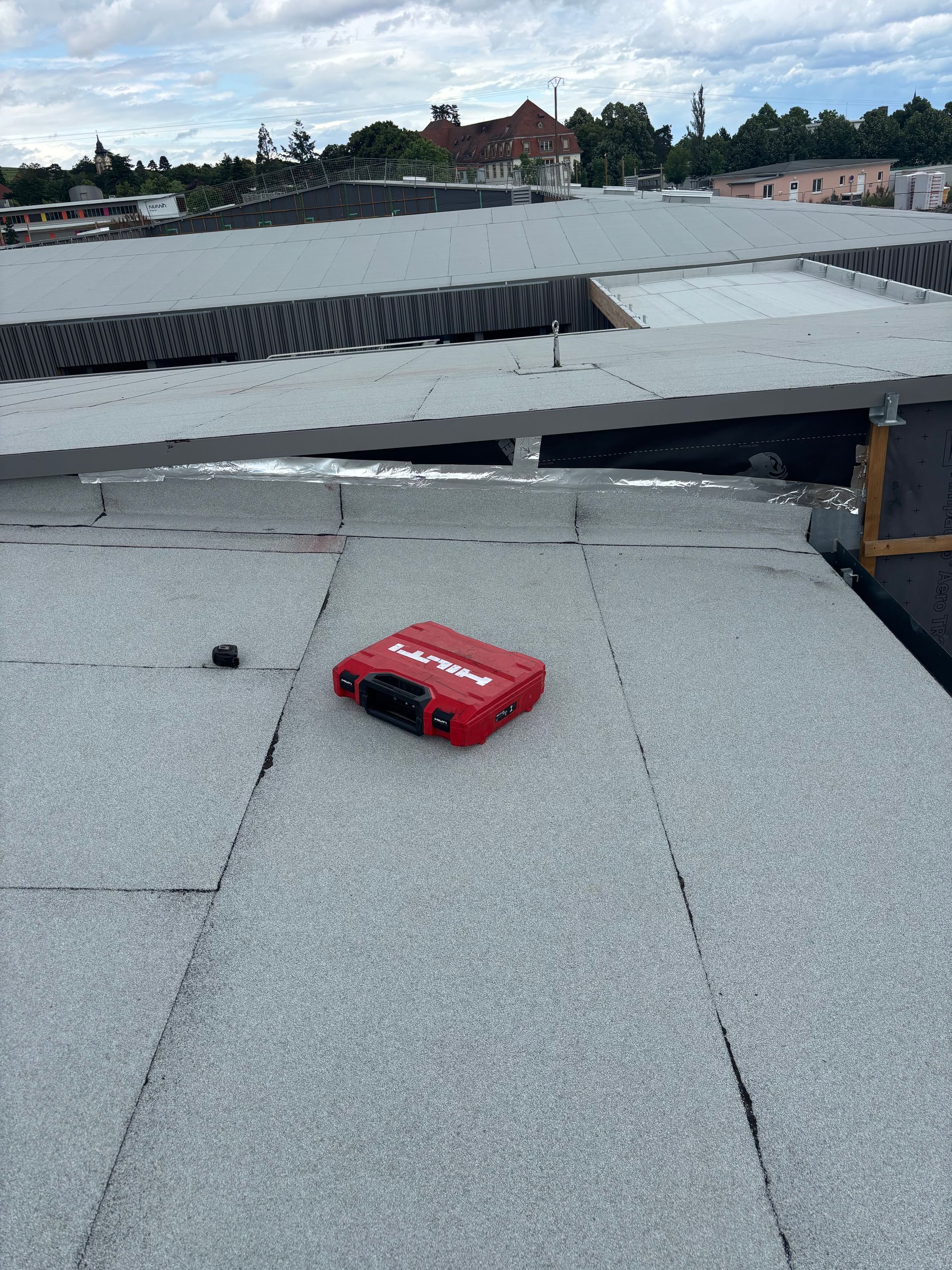 Une boîte à outils rouge sur un toit plat recouvert de papier goudronné gris, avec un paysage urbain au loin.