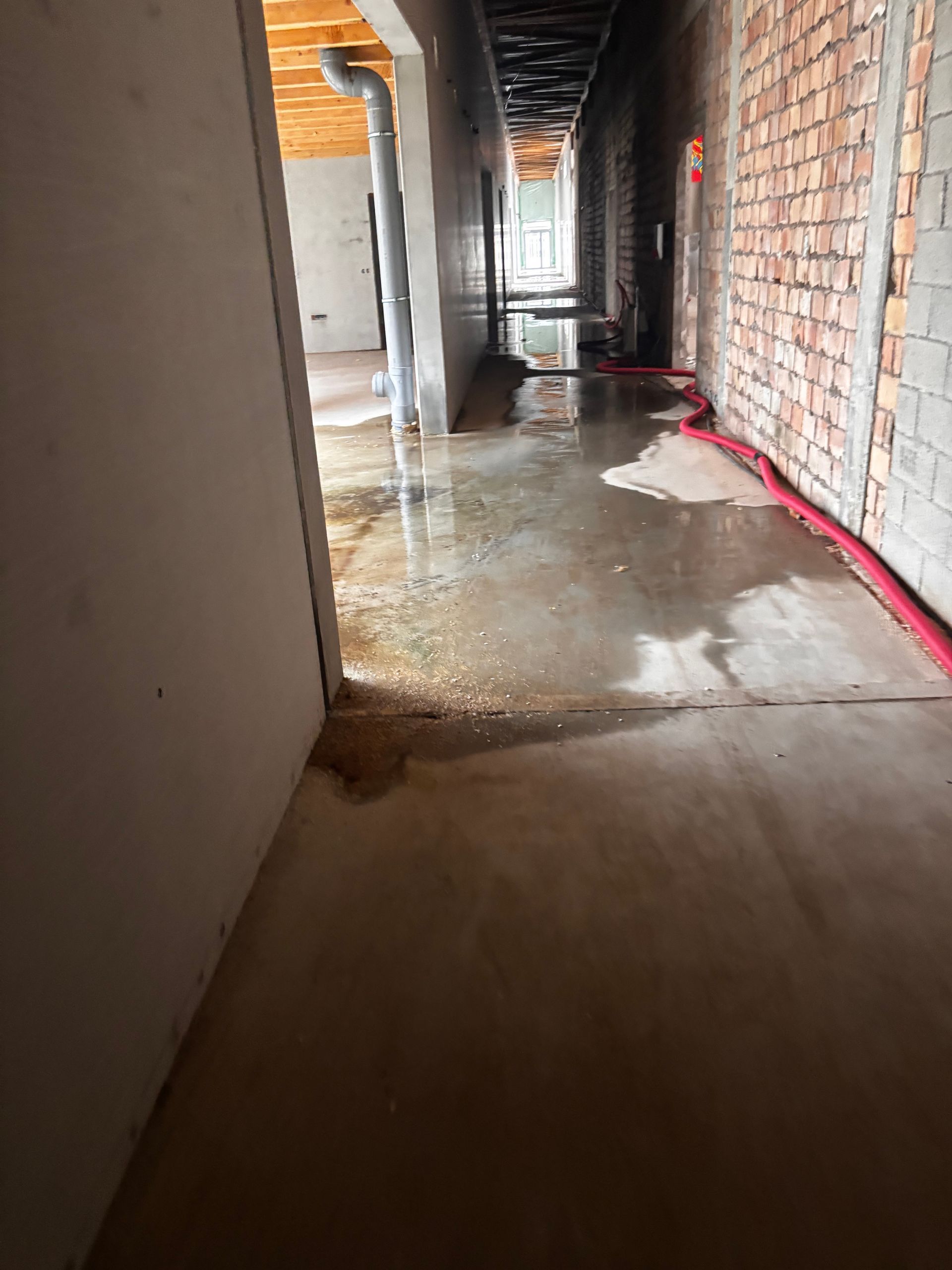 Couloir de chantier avec sol en béton frais, murs en briques et plâtre non finis et tuyaux apparents.