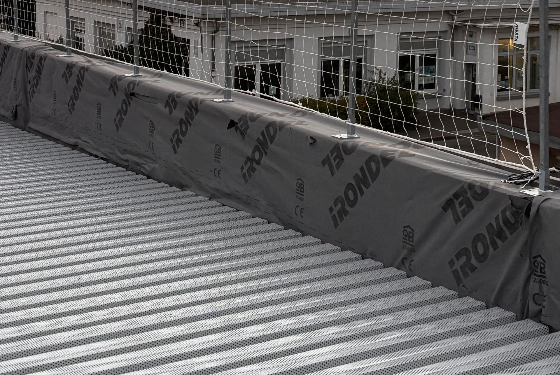 Toiture métallique recouverte d'une bâche et d'un filet de protection noirs. Bâtiment en arrière-plan.