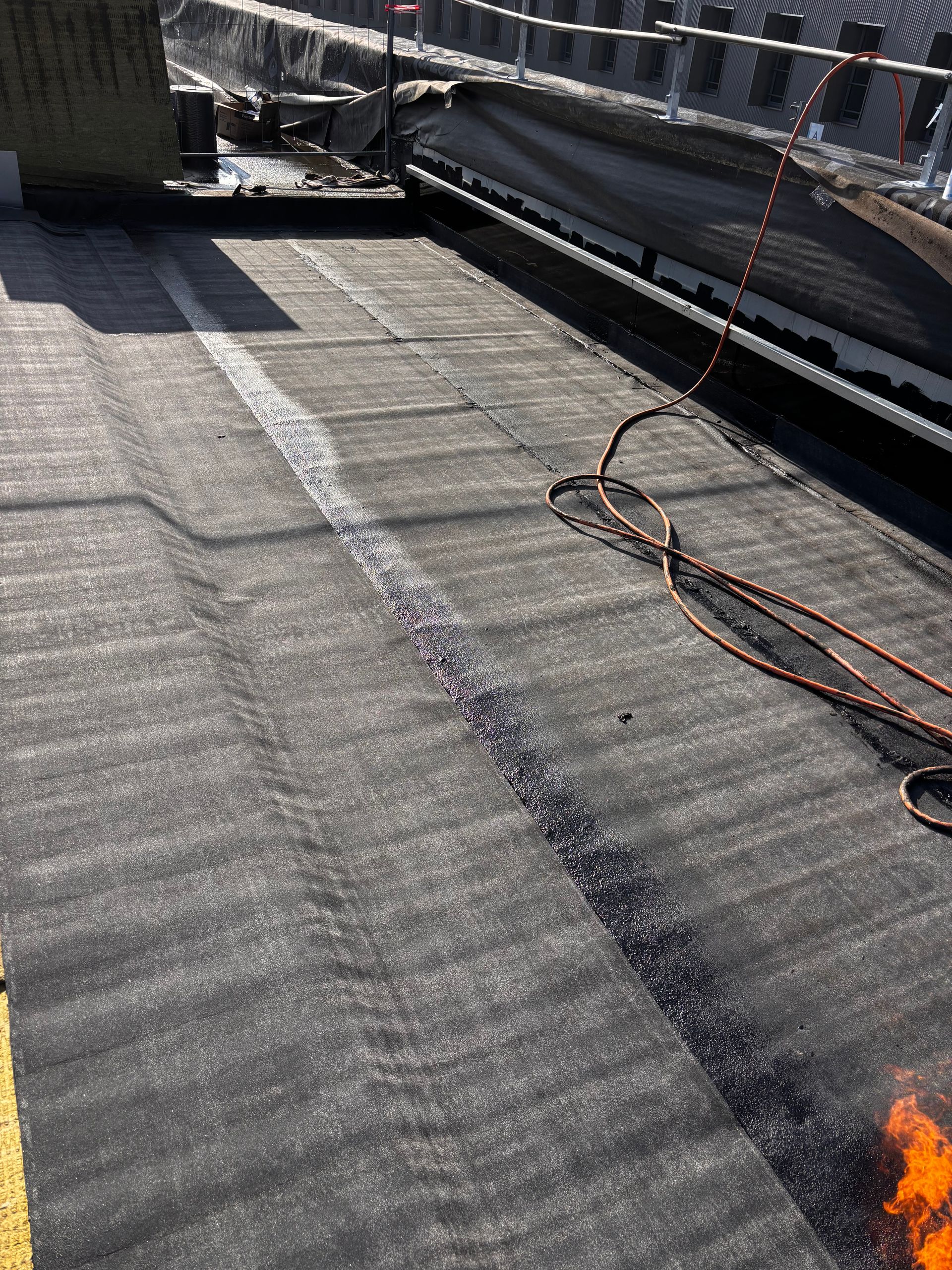 Application d'un matériau de toiture noir ; une flamme est visible dans le coin inférieur droit.