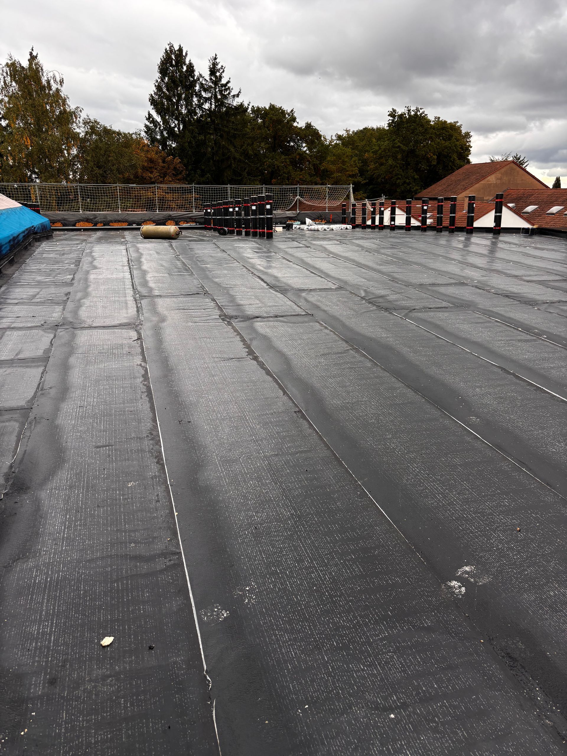 Toit plat noir avec des lignes de joint blanches et quelques équipements, sous un ciel couvert.