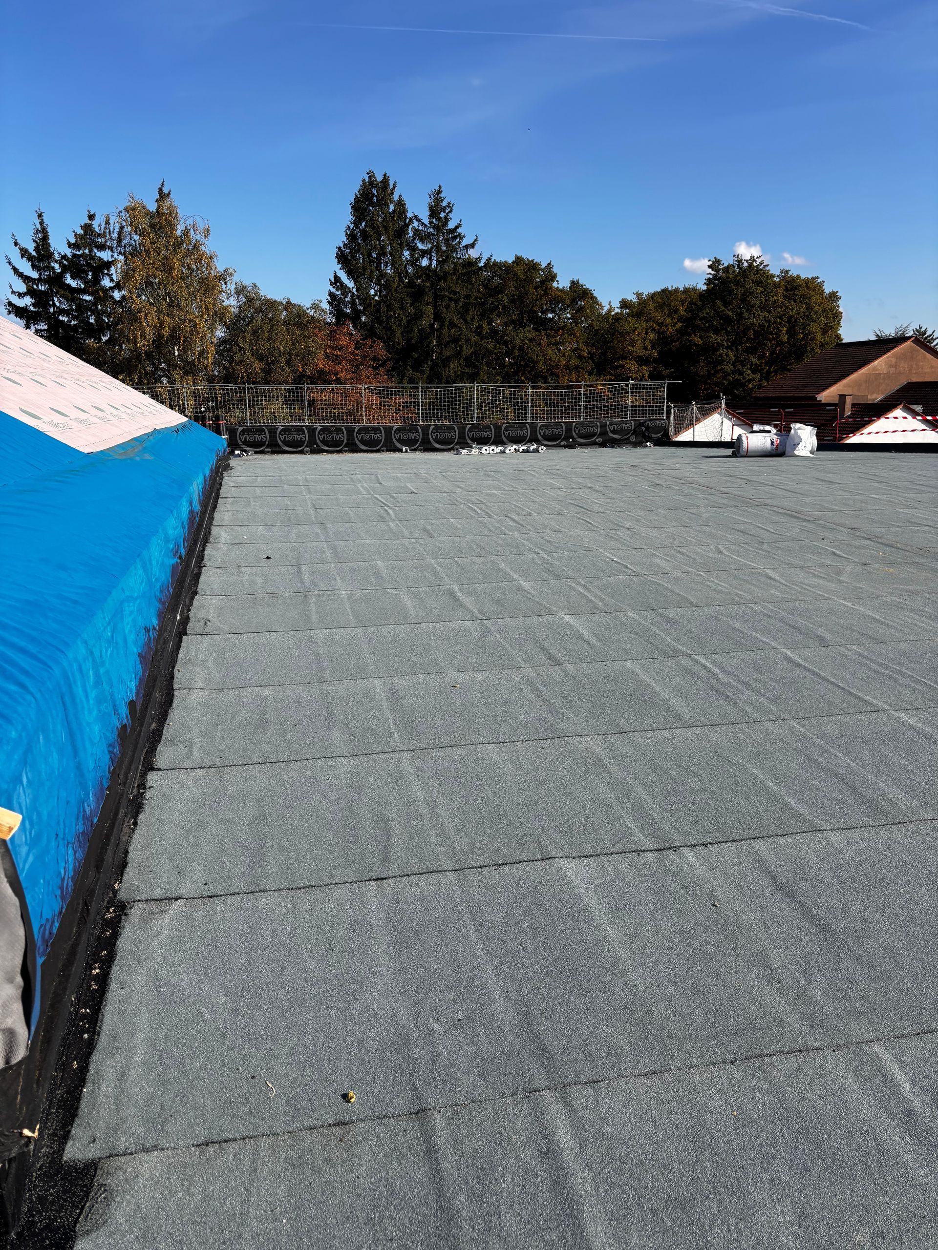 Toit plat à surface gris foncé, bordure bleue, arbres et ciel bleu.