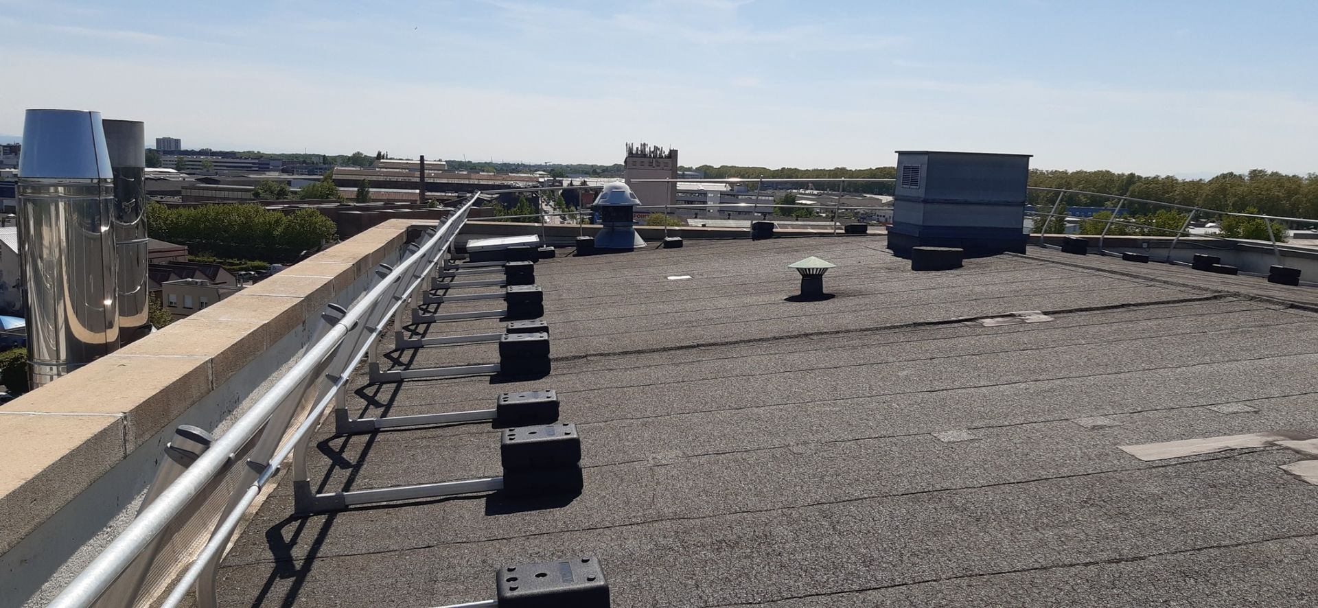 Vue depuis le toit d'un immeuble, avec des tuyauteries en bordure, divers équipements et la ville en arrière-plan.