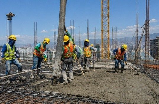 Obreros de la construcción con cascos y chalecos vertiendo hormigón sobre una base de barras de refuerzo al aire libre.