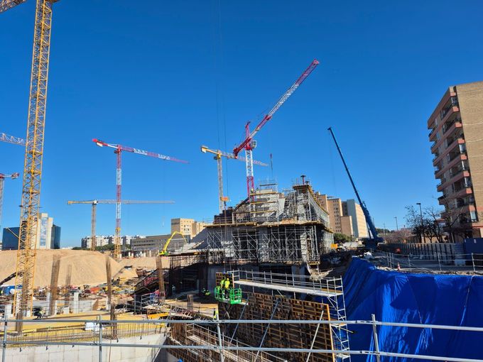 Un trabajador con casco se encuentra debajo de un puente en construcción con andamios y grúas cerca de un río.