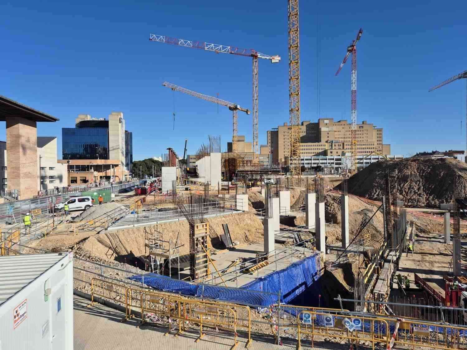 Obra en construcción con grúas, estructura de acero y edificios de apartamentos circundantes bajo un cielo azul despejado.