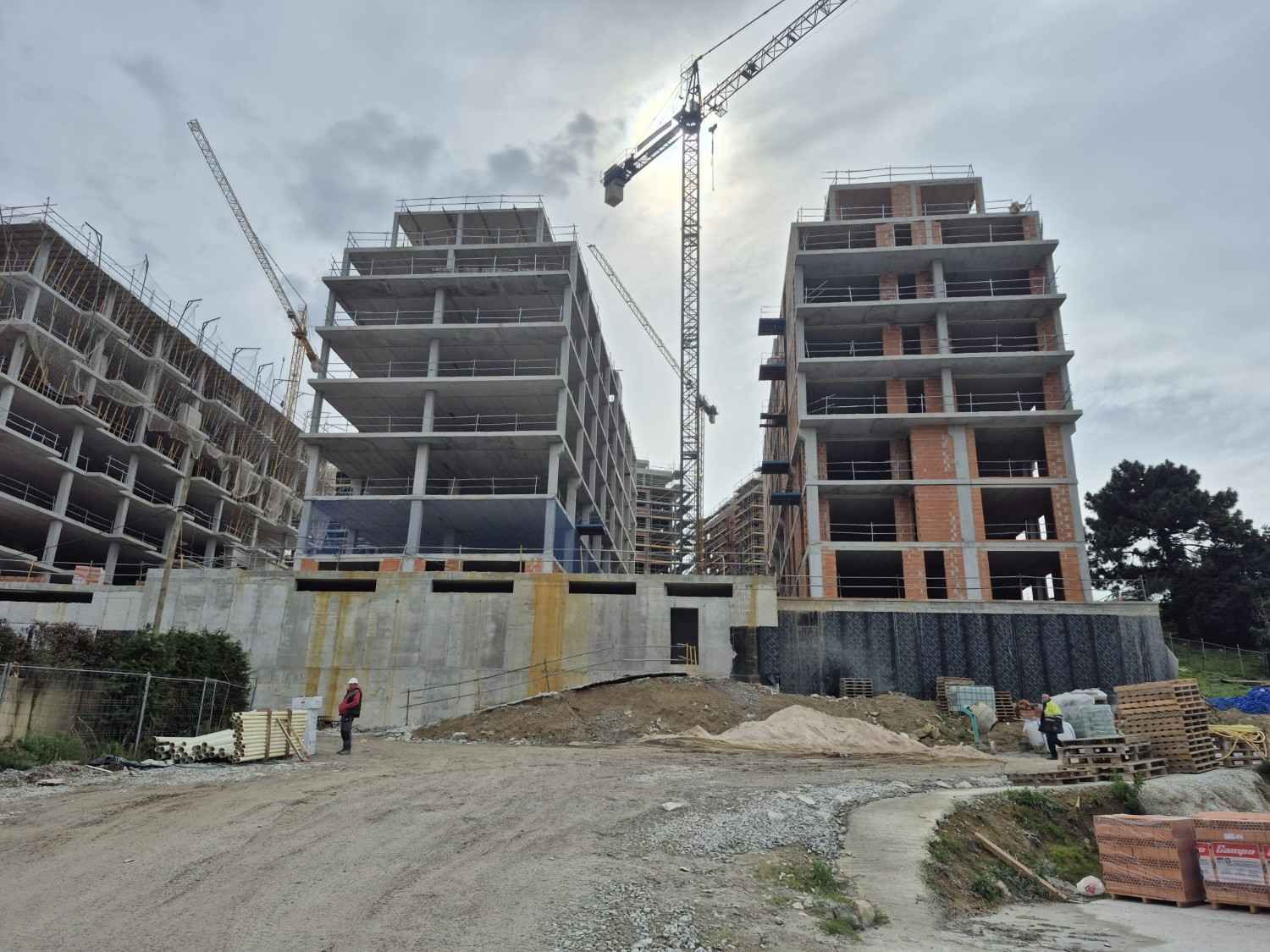 Obra en construcción con dos edificios de varias plantas sin terminar, grúas y un camino de tierra en primer plano.