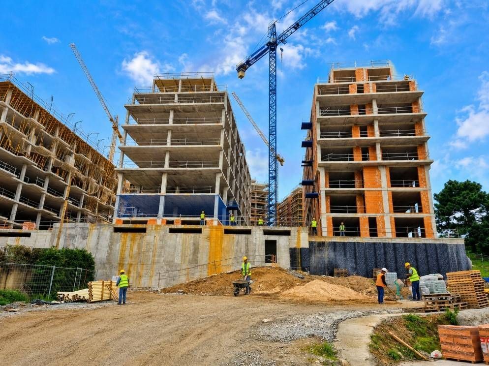 Obra en construcción con edificios de apartamentos sin terminar, grúas y trabajadores bajo un cielo azul.