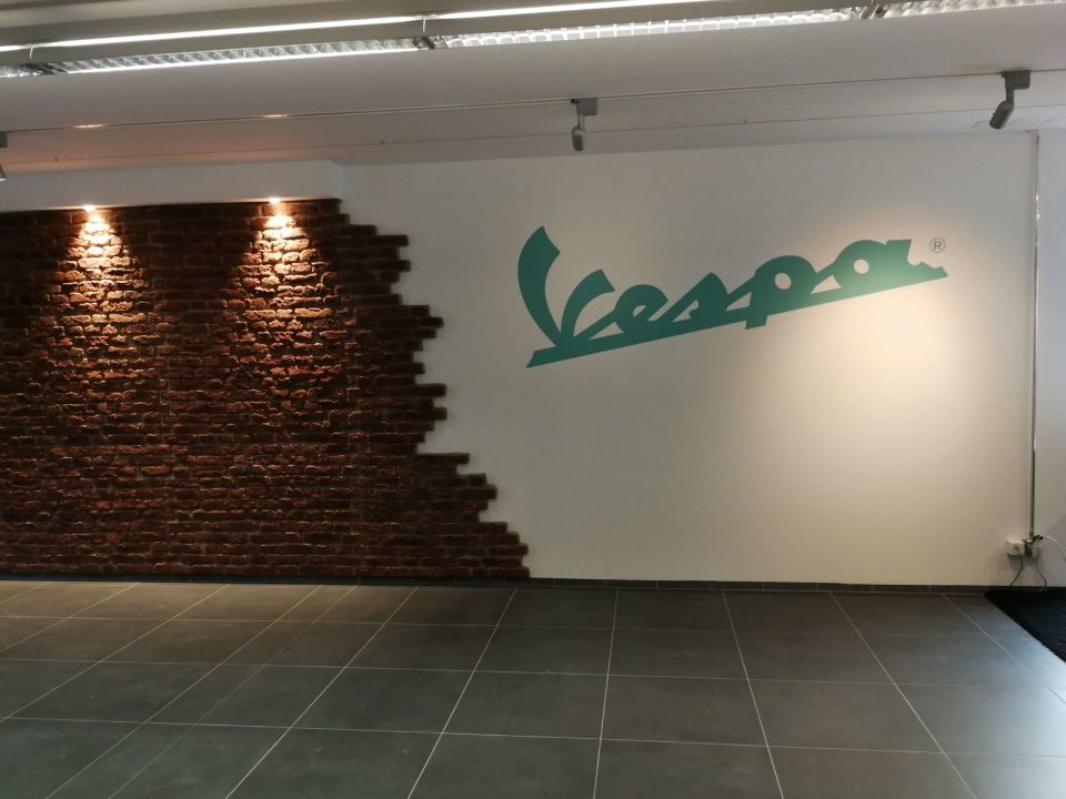 Weisse Wand mit Vespa-Logo, teilweise gemauerter Wandabschnitt und grauer Fliesenboden.