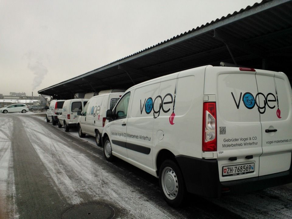 Weisse Vogel-Lieferwagen parkten unter einem überdachten Gebäude; der Boden war schneebedeckt, der Himmel grau, und in der Ferne war eine Fabrik zu sehen.