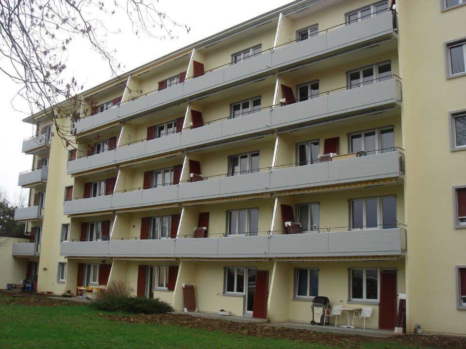 Gelbes Mehrfamilienhaus mit mehreren Etagen und Balkonen. Rote Türen und Fensterläden.