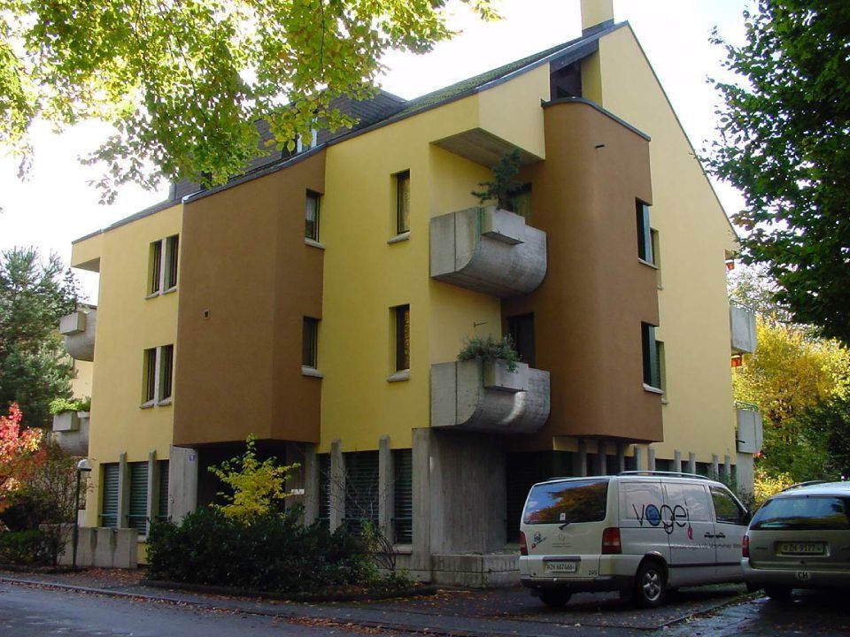 Dreistöckiges, gelb-braunes Gebäude mit Betonbalkonen, im Hintergrund Bäume, davor ein Lieferwagen und ein Auto geparkt.