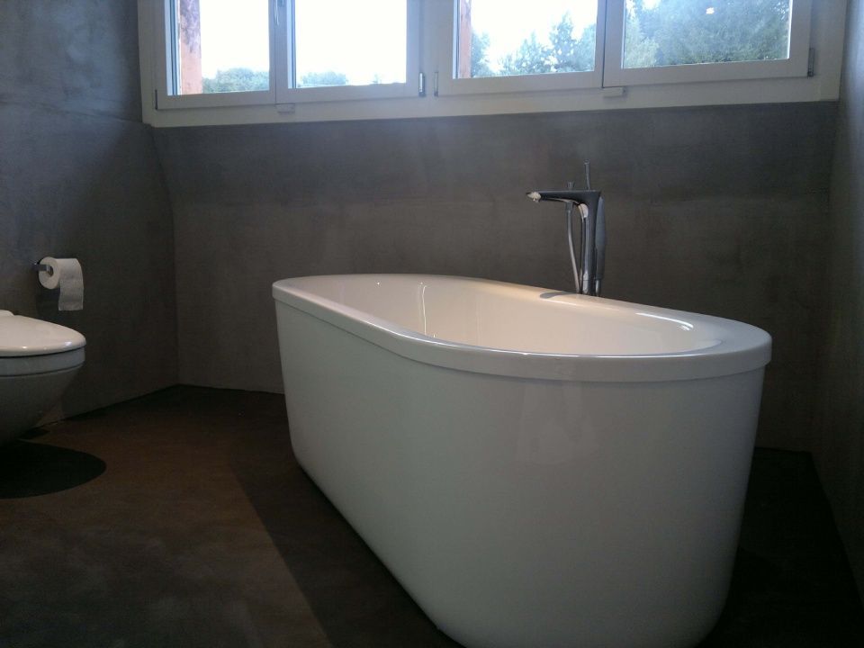 Badezimmer mit weisser Badewanne, Toilette und moderner Armatur. Graue Wände und dunkler Boden. Fenster im Hintergrund.