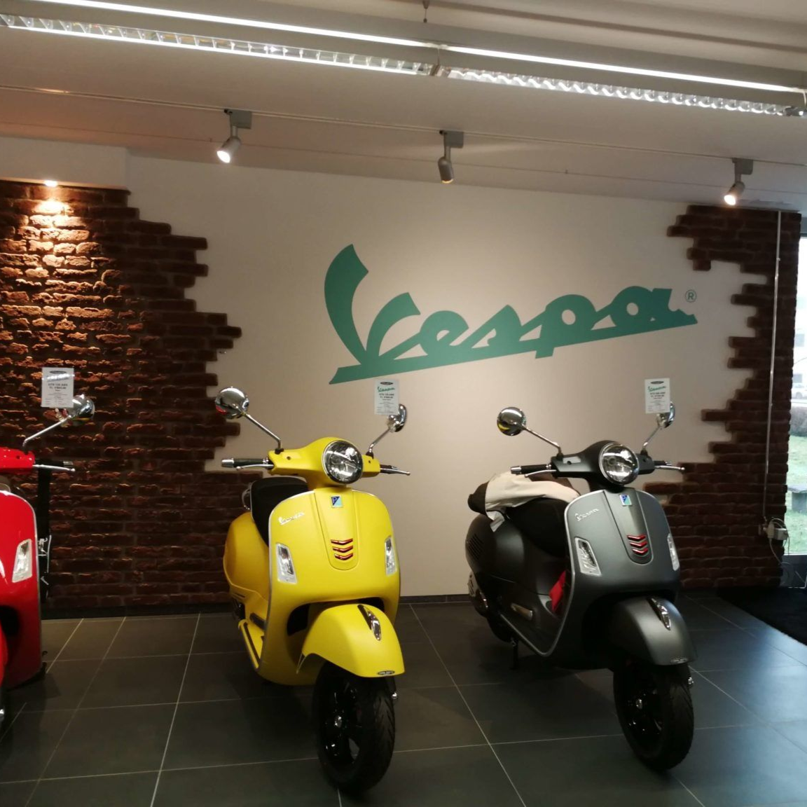 Weisse Wand mit Vespa-Logo, teilweise gemauerter Wandabschnitt und grauer Fliesenboden.