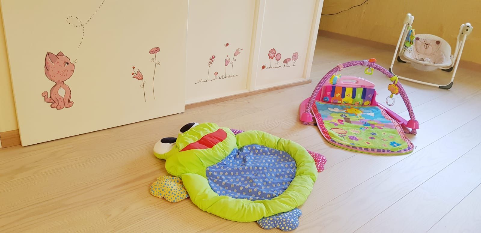 Una alfombra de juego con forma de rana se encuentra sobre un piso de madera al lado de una cuna.