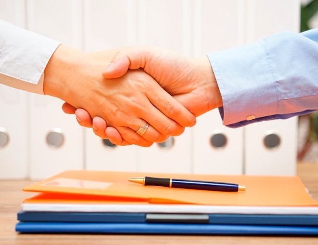 Dos personas se dan la mano sobre una mesa con carpetas y un bolígrafo.