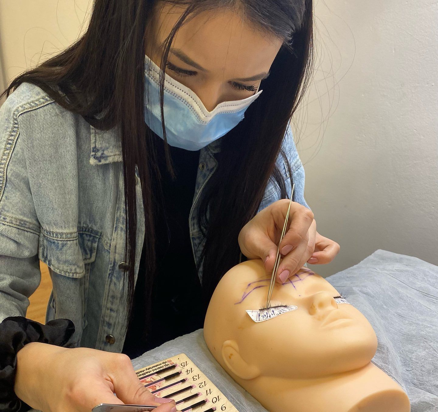 Una persona con una mascarilla azul practica la aplicación de extensiones de pestañas en la cabeza de un maniquí 
