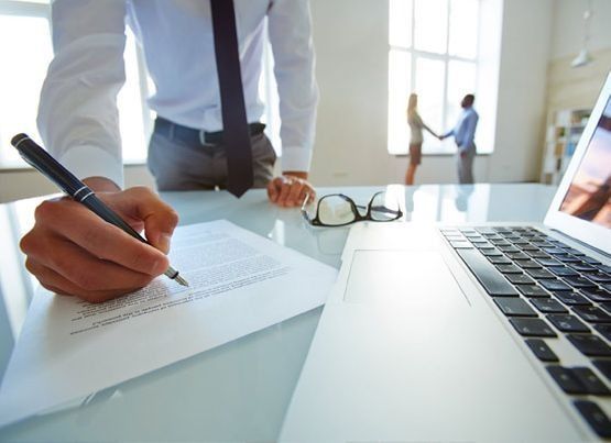 Persona escribiendo en papel, computadora portátil y dos personas dándose la mano en el fondo junto a la ventana.