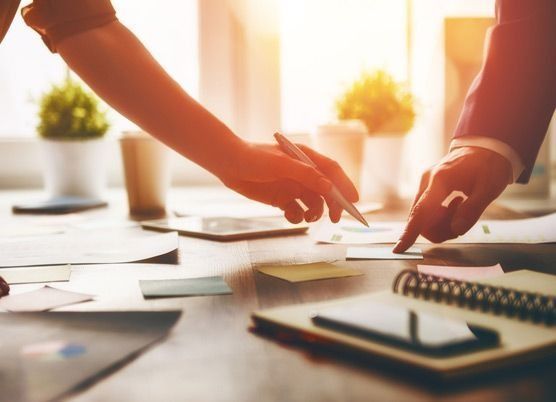Dos personas en un escritorio, señalando y escribiendo en papeles. La luz del sol entra a raudales.