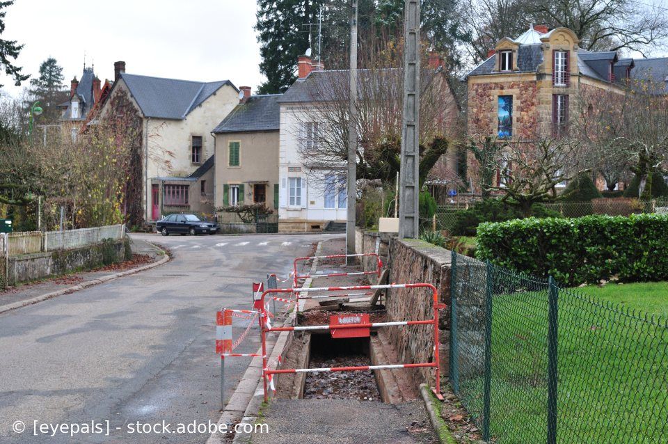 Straße mit im Bau befindlichem Graben, umgeben von Häusern und Baustellenabsperrungen.