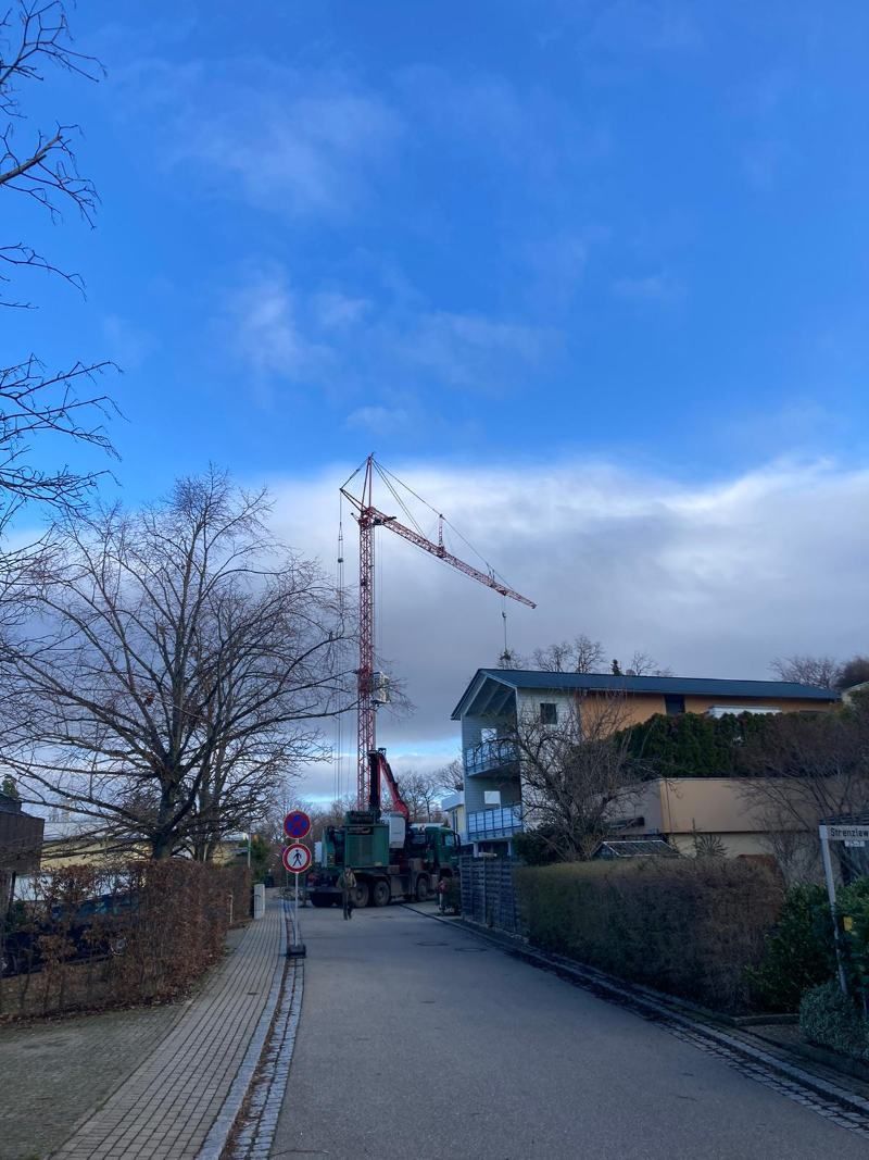 Ein hoher roter Baukran über einer Straße mit Häusern und einem bewölkten blauen Himmel.
