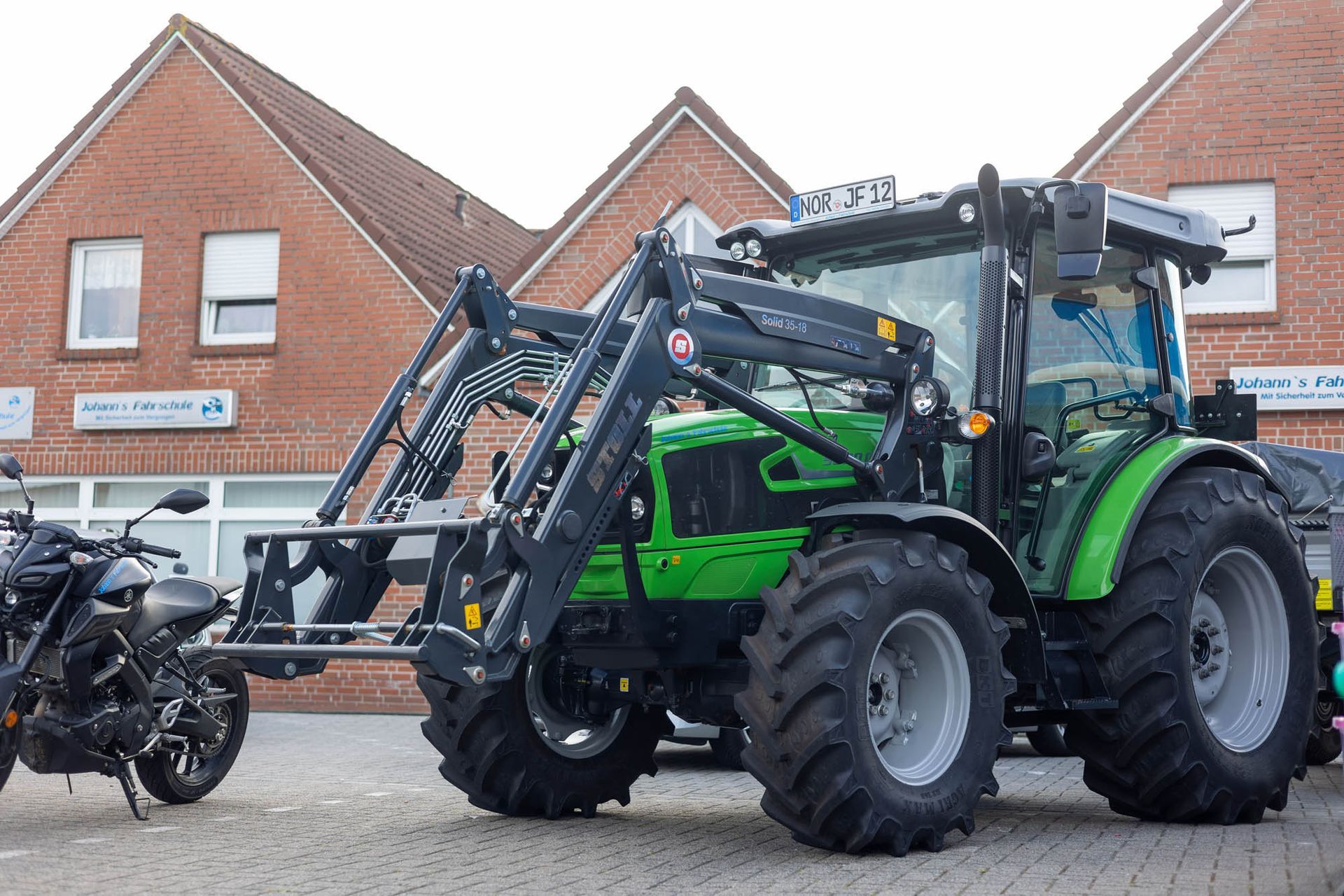 Ein grüner Traktor parkt neben einem Motorrad vor einem Backsteingebäude.