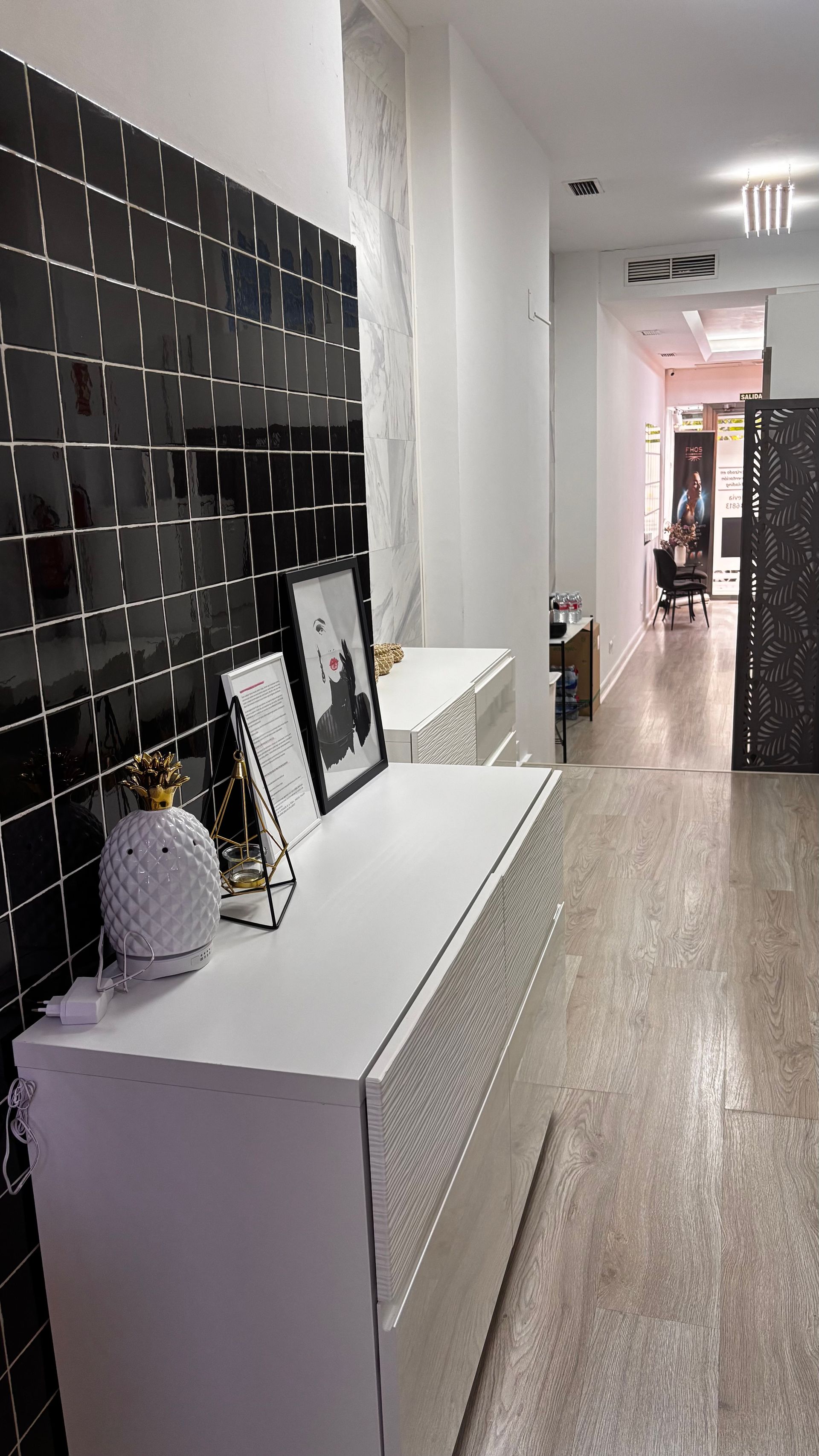Cómoda blanca con decoración, pared de azulejos negros, pasillo con luz y mampara negra.